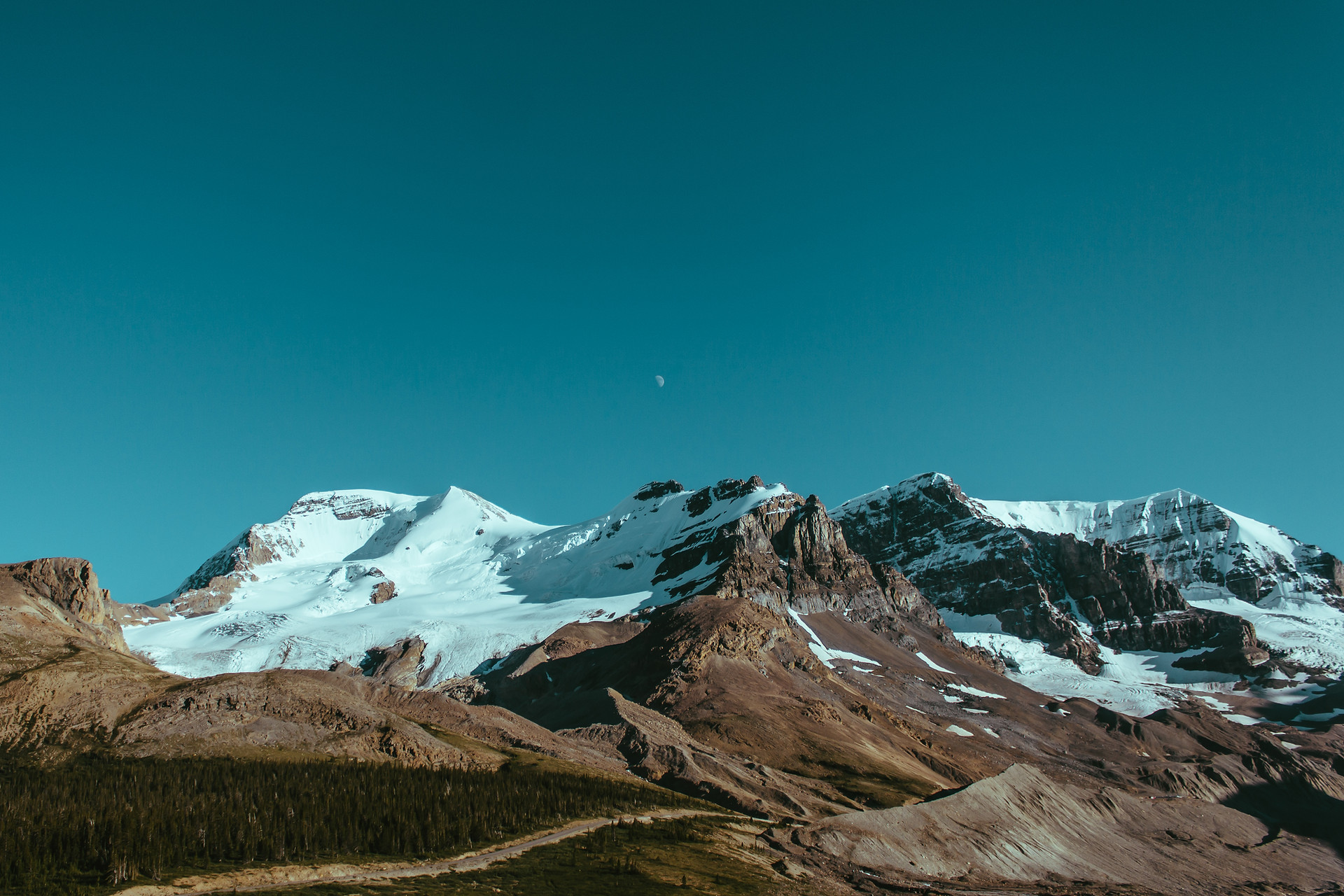 Snow-capped mountain range under a deep teal sky with a faint moon, forested foothills below.
