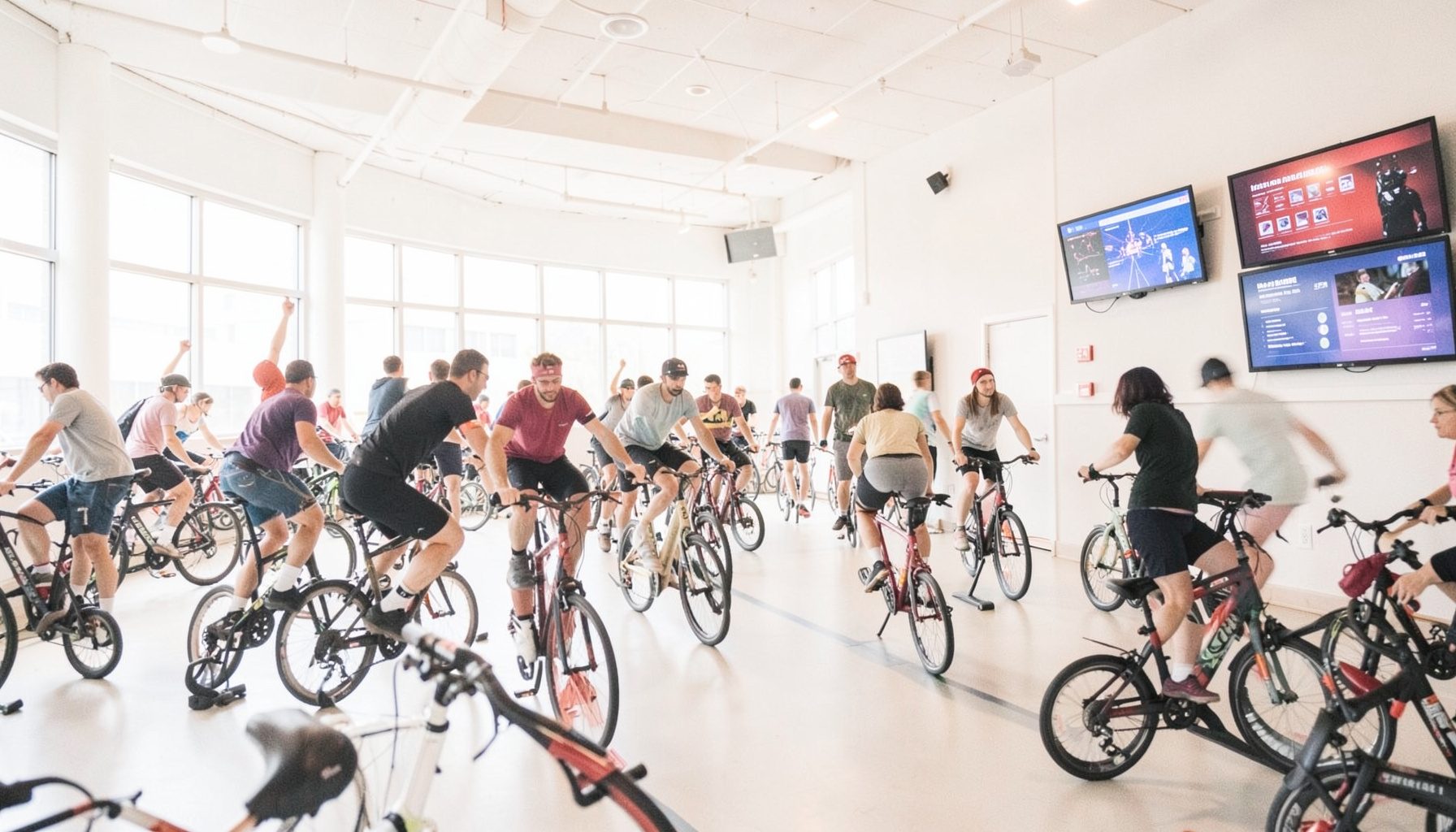 A wide shot of an indoor cycling event space with multiple riders, screens mounted on the wall, energetic group atmosphere