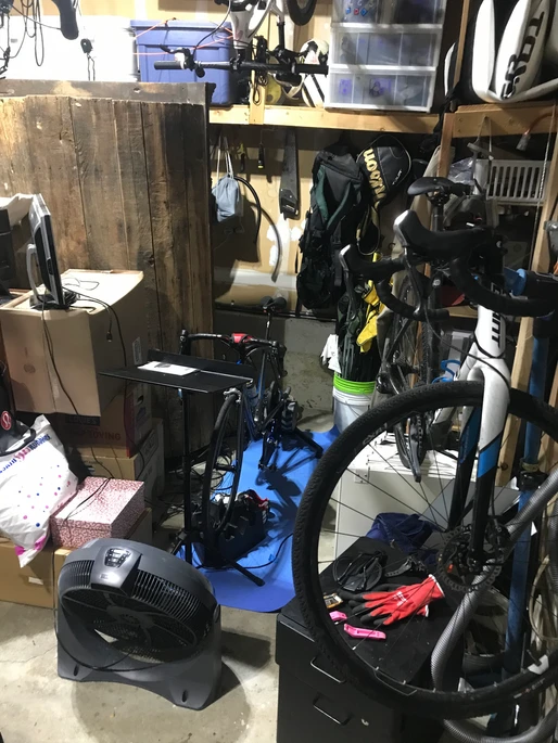 Garage storage area with bicycles, a fan, plastic bins and shelving, dim cool lighting.